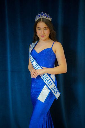 A pageant contestant wearing a sparkling royal blue evening gown and tiara poses against a dark blue backdrop.