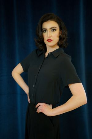 A stylish retro-inspired portrait against a blue backdrop featuring a classic black button-up dress with short sleeves.