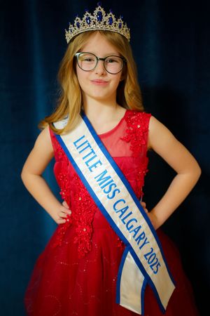 A beauty pageant contestant wearing glasses and a sparkly red dress poses with a crown and "Little Miss Calgary" sash.