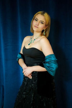Elegant portrait of a woman in black evening gown with teal wrap and statement necklace against dark blue backdrop.