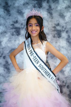 Young pageant contestant wearing white tulle gown and sparkly tiara poses against cloudy backdrop.