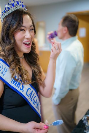 A pageant winner wearing a blue sash and sparkly tiara smiles joyfully in a black dress while holding something in her hands.