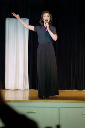 A performer in a long black dress stands on stage with one arm raised while singing into a microphone.