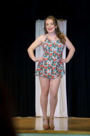 A person poses on stage wearing a floral print romper and wedge sandals during what appears to be a fashion show.