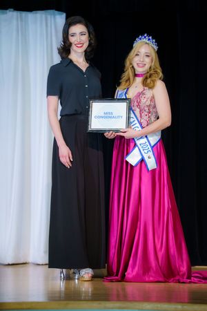 Two people stand on stage at a formal event, one wearing a dark button-up outfit and another in a sparkly pink evening gown.