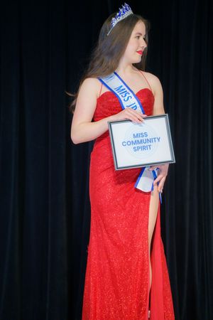 A pageant contestant in a red evening gown holds a 'Miss Community Spirit' sign while wearing a tiara on stage.