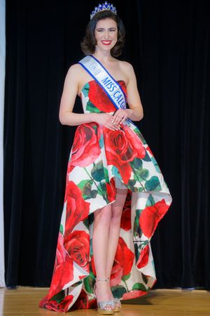 Beauty pageant contestant wearing crown and floral high-low dress with red rose pattern poses on stage.
