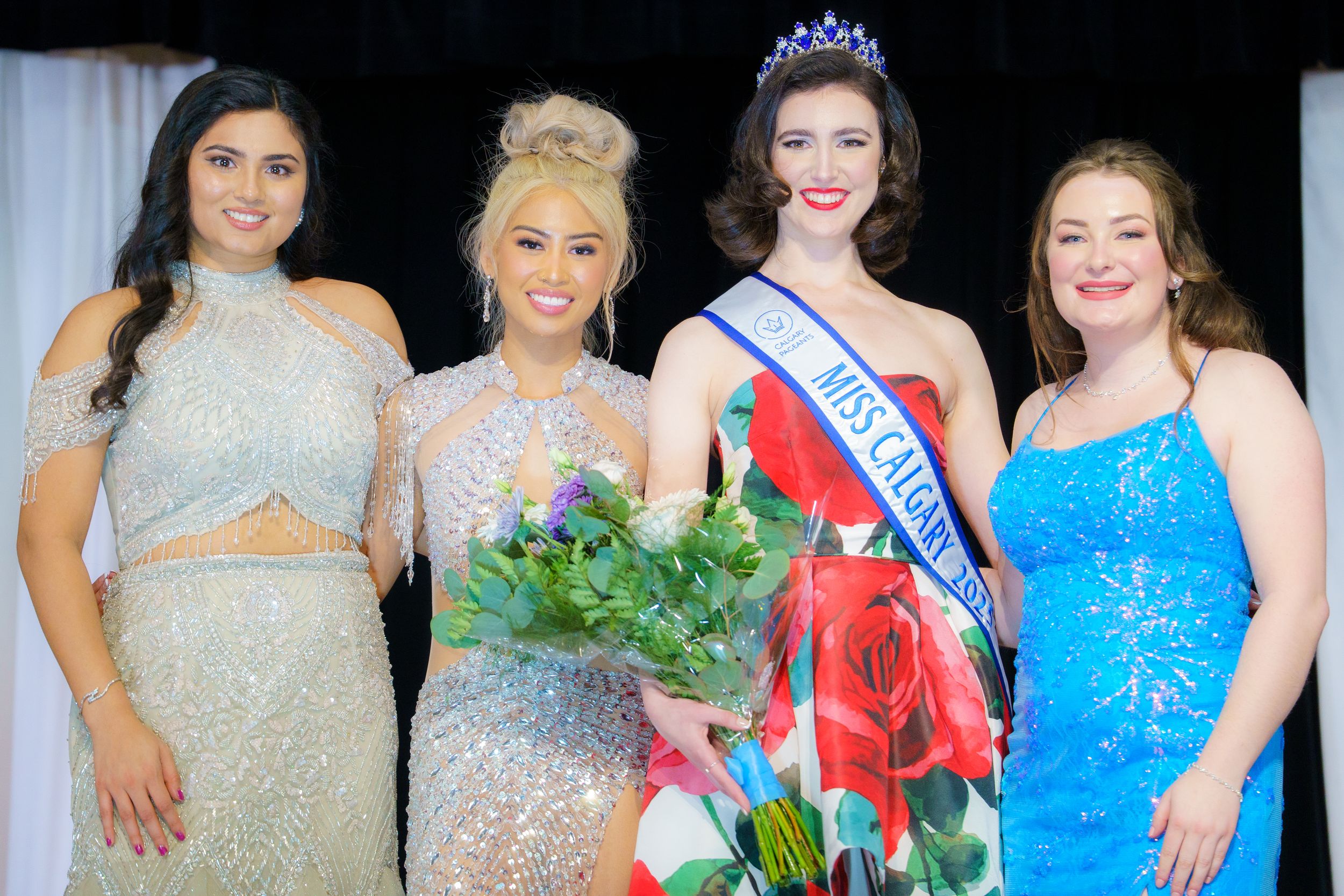 Four people pose together at a formal event wearing sparkly evening gowns in silver and blue tones.