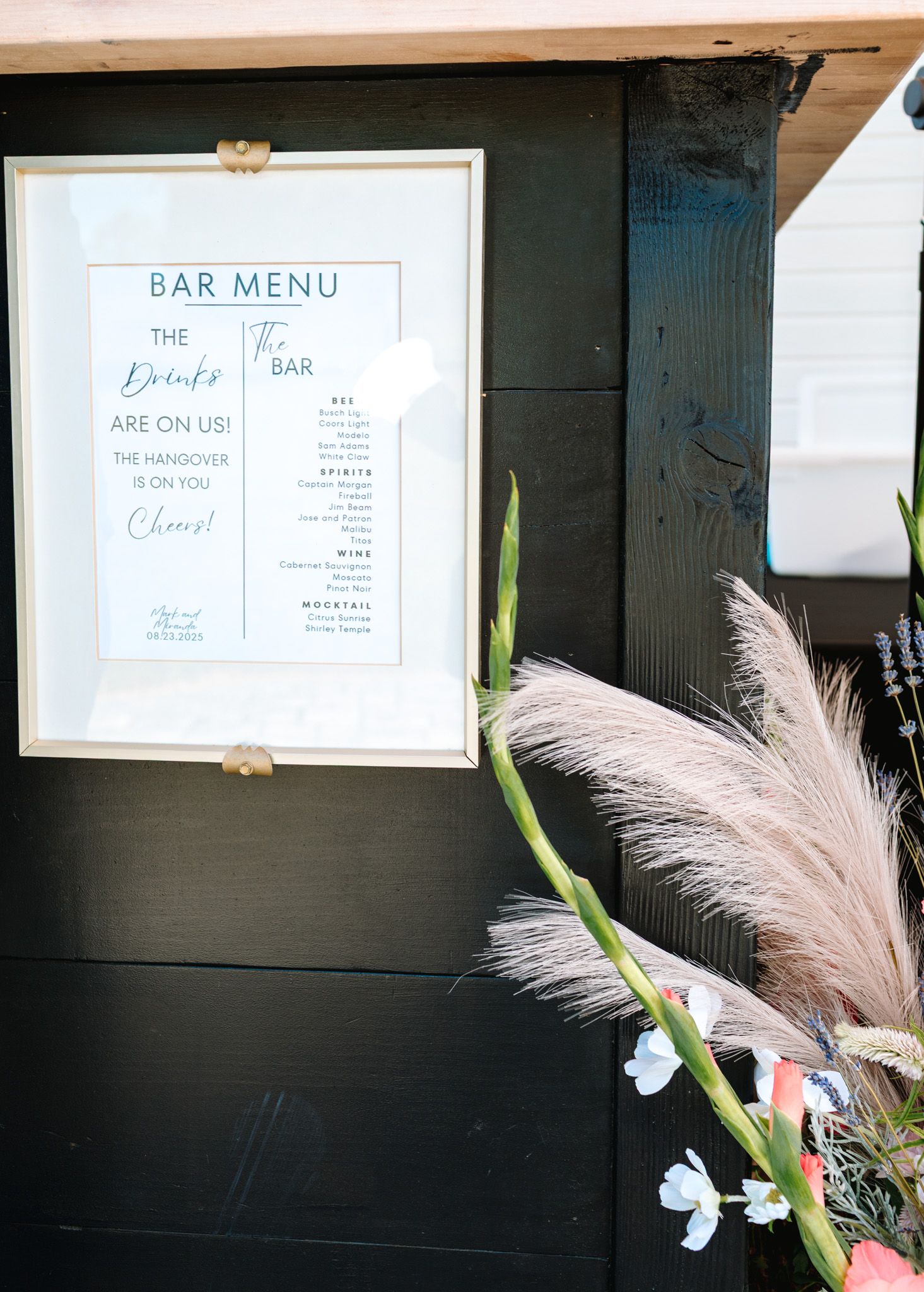 Framed bar menu sign on black wood wall with pampas grass and floral decorations nearby.