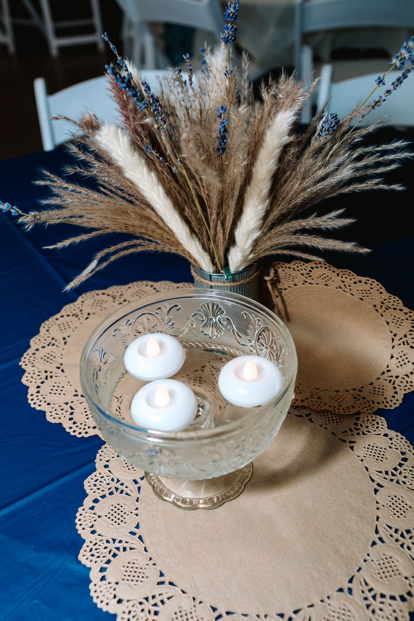 Rustic centerpiece with pampas grass, lavender, and floating candles in glass bowl on lace doily.
