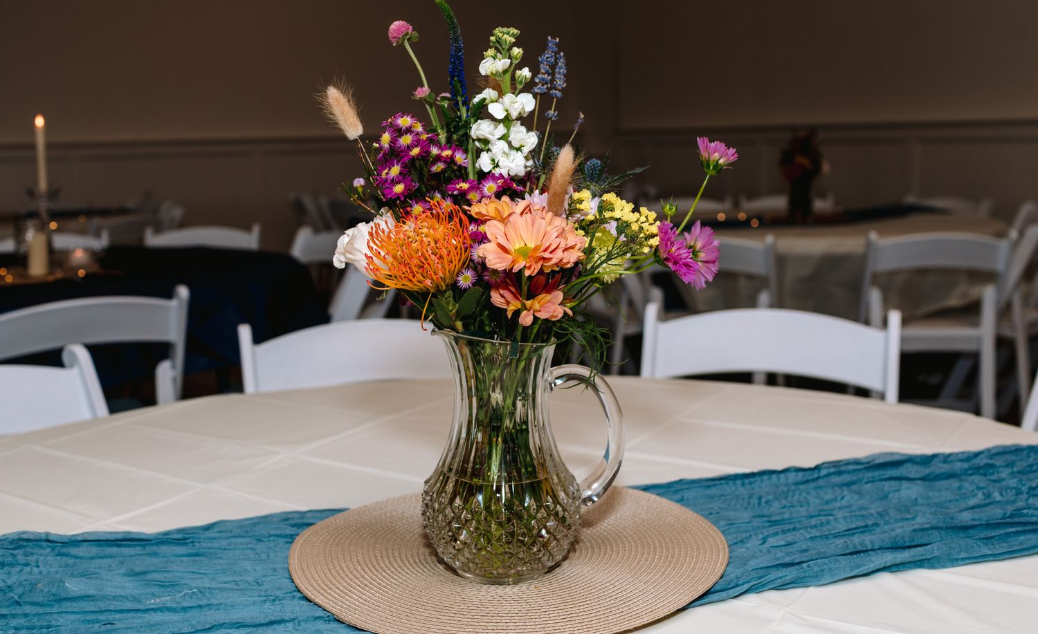 Colorful wildflower bouquet in glass pitcher centerpiece on teal table runner at wedding reception.