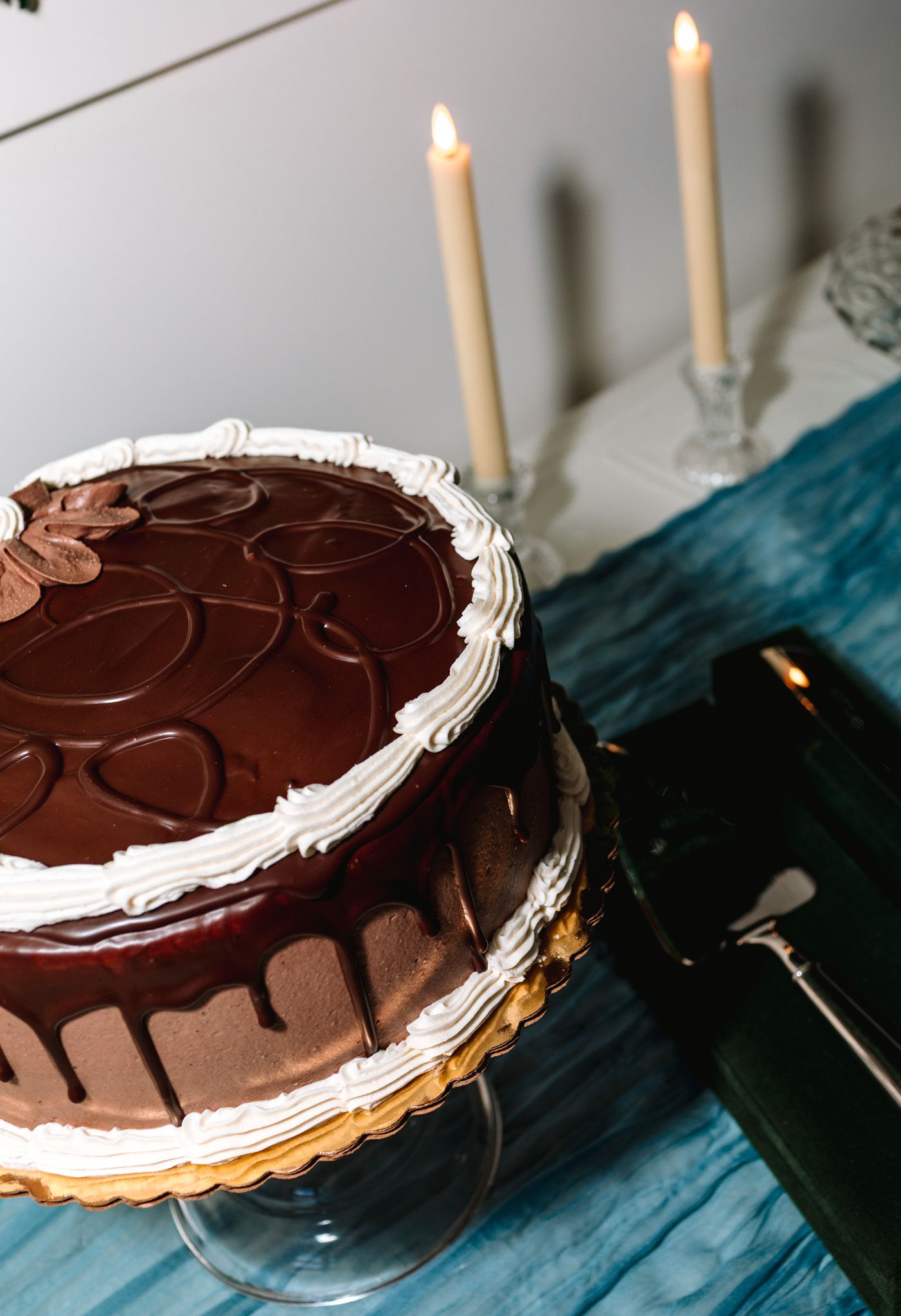 Decadent chocolate drip wedding cake with white frosting and candles on blue tablecloth.