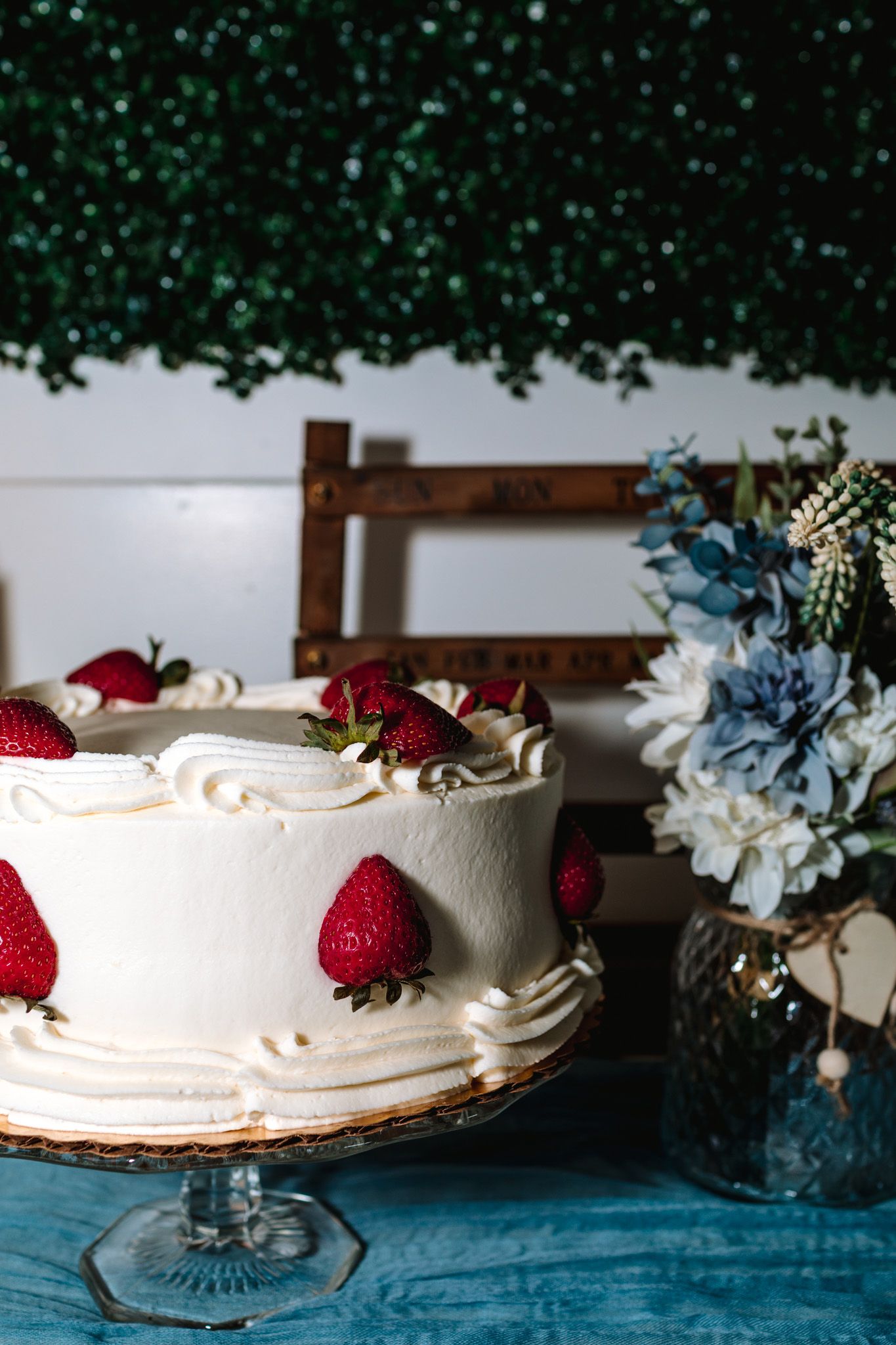 White wedding cake topped with fresh strawberries on glass stand with blue floral arrangement.