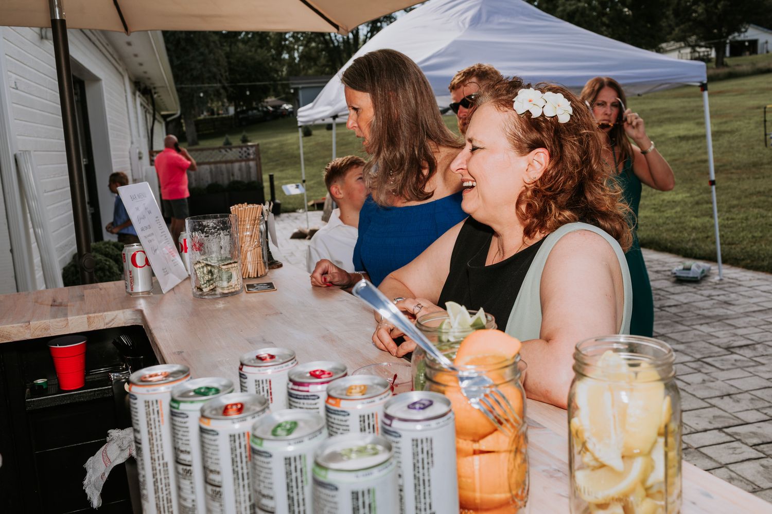 Women smiling at an outdoor bar with canned drinks and mason jars on a sunny day.