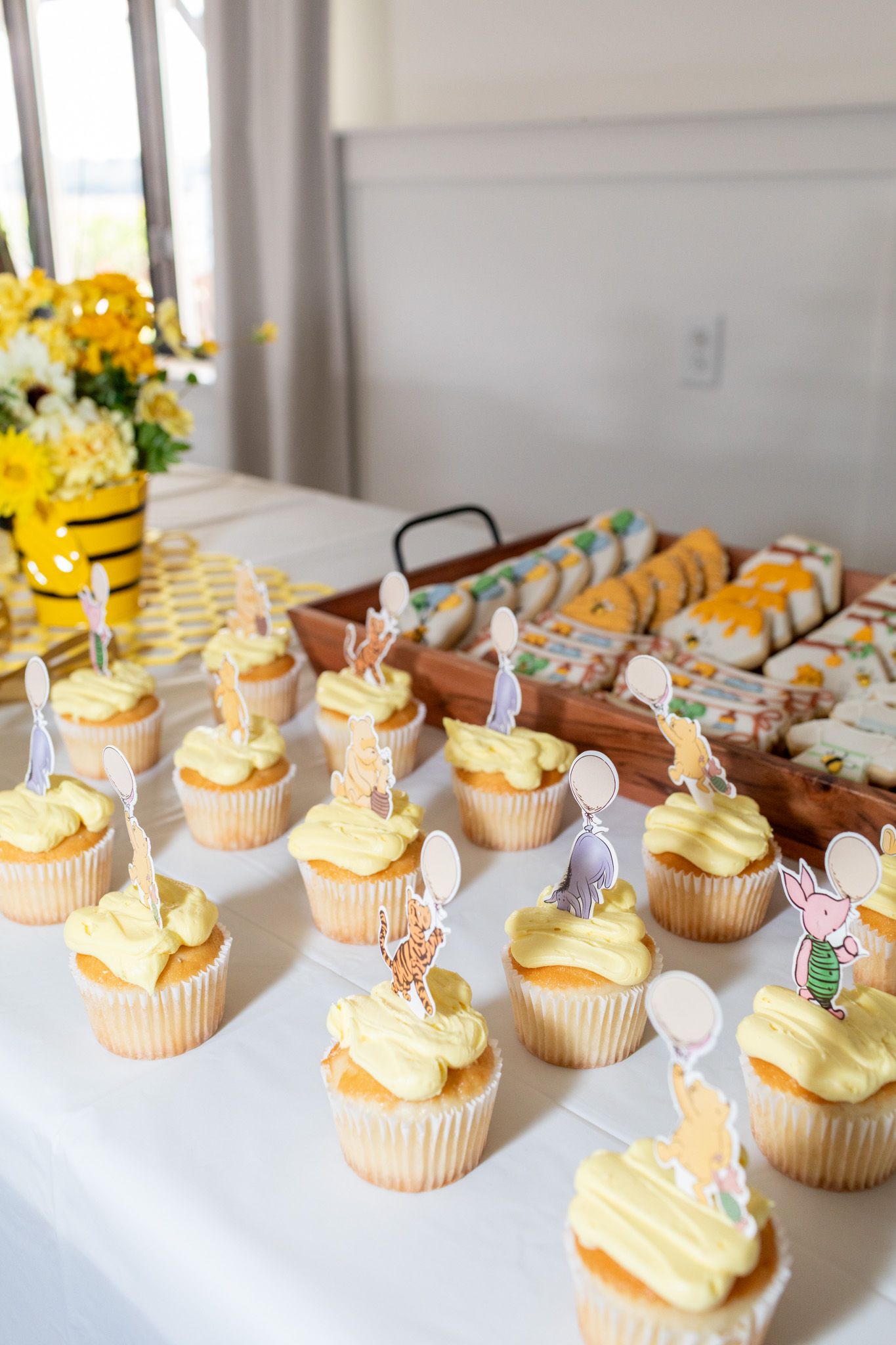 Yellow frosted cupcakes with Winnie the Pooh character toppers displayed on a white table at a baby shower.