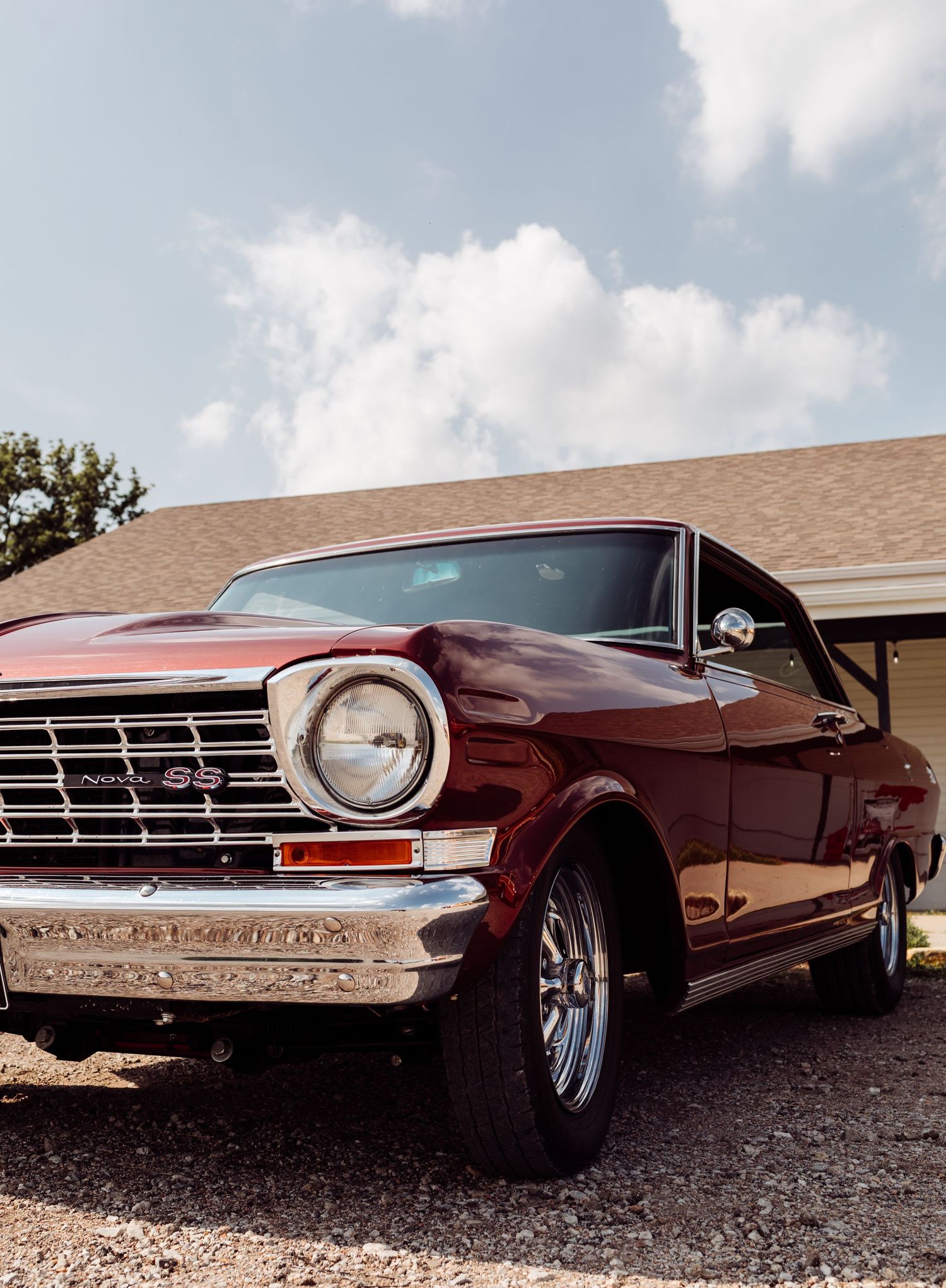 A restored burgundy Chevrolet Nova SS muscle car with chrome grille parked in a driveway under a partly cloudy sky.