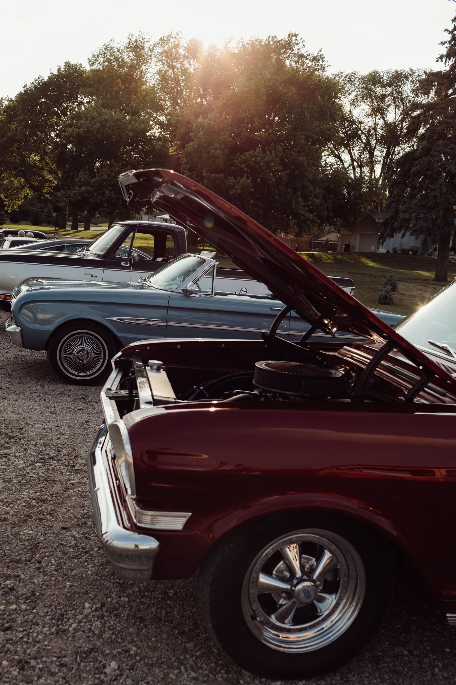 A dark red classic car with hood open at a vintage car show, with a blue convertible parked behind at sunset.