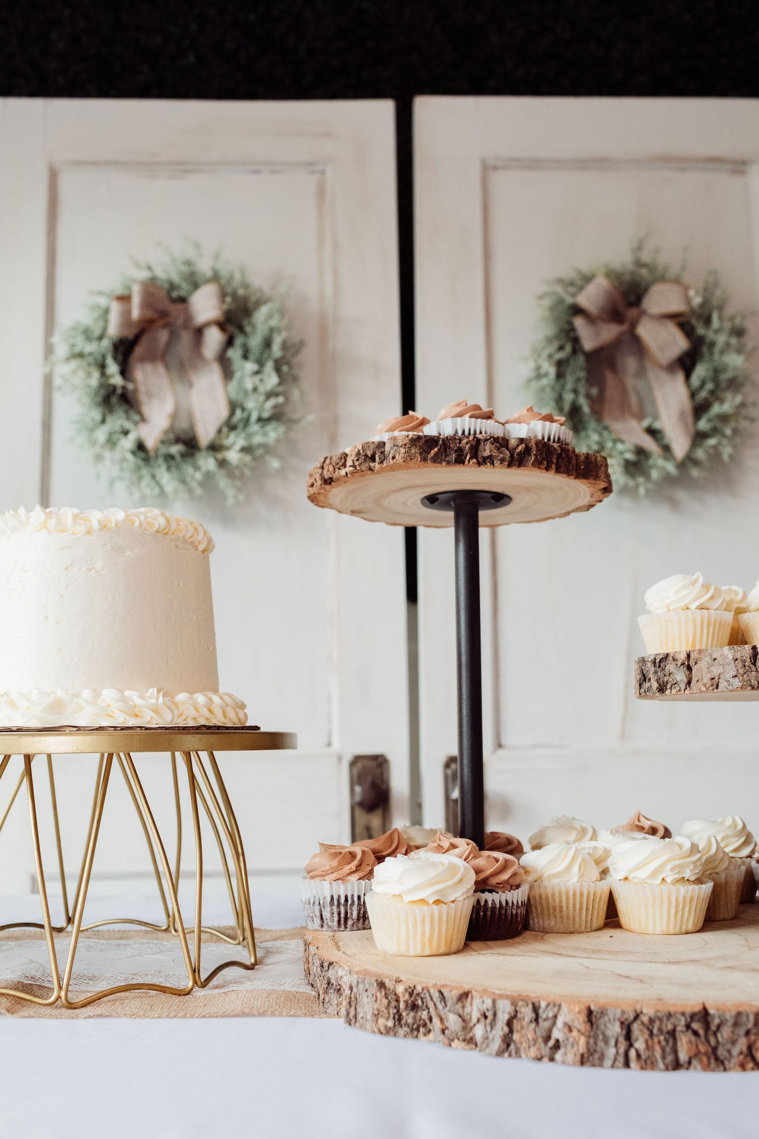 Rustic wedding dessert table with white cake on gold stand and cupcakes on wood slice displays near wreath doors.