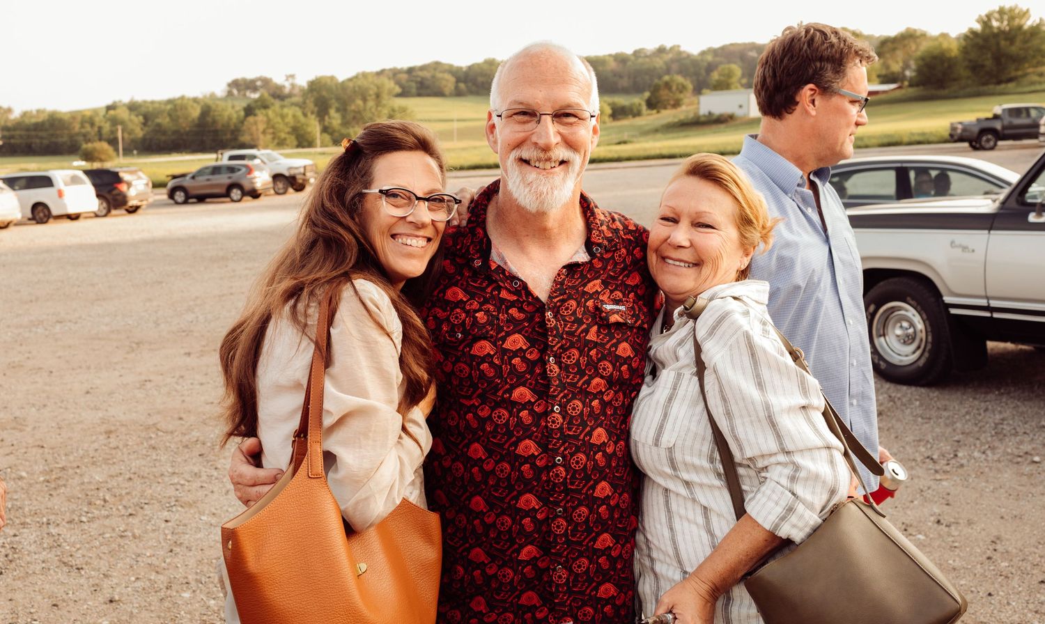 Three smiling people posing together outdoors at a classic car show event during golden hour evening light.
