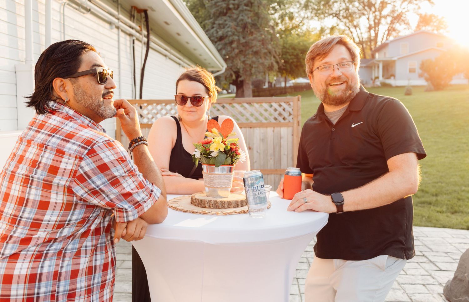 Three friends socializing at an outdoor gathering around a cocktail table with flowers in warm evening sunlight.