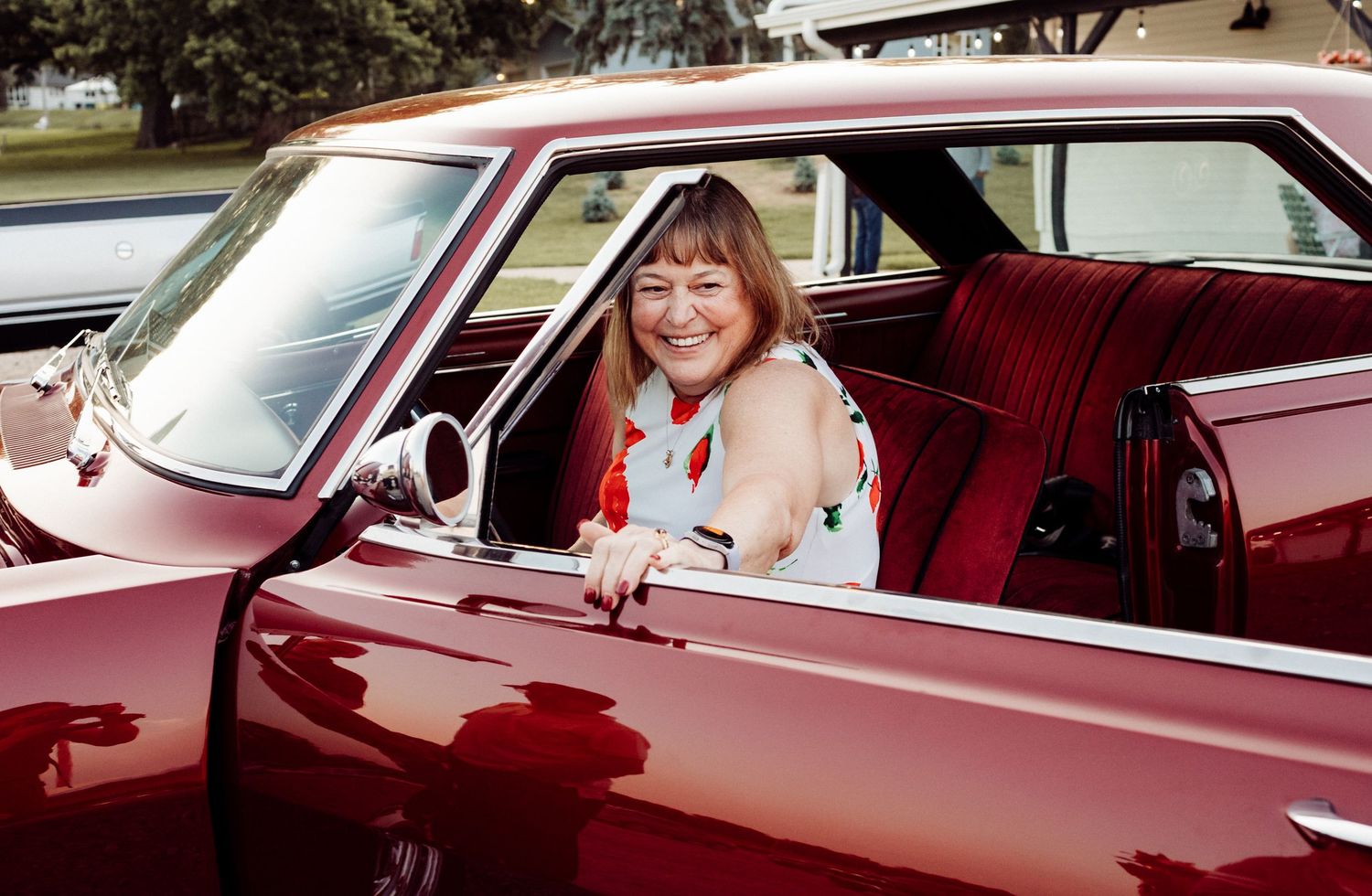 A smiling woman sitting in the red interior of a classic vintage car with the door open on a sunny day.