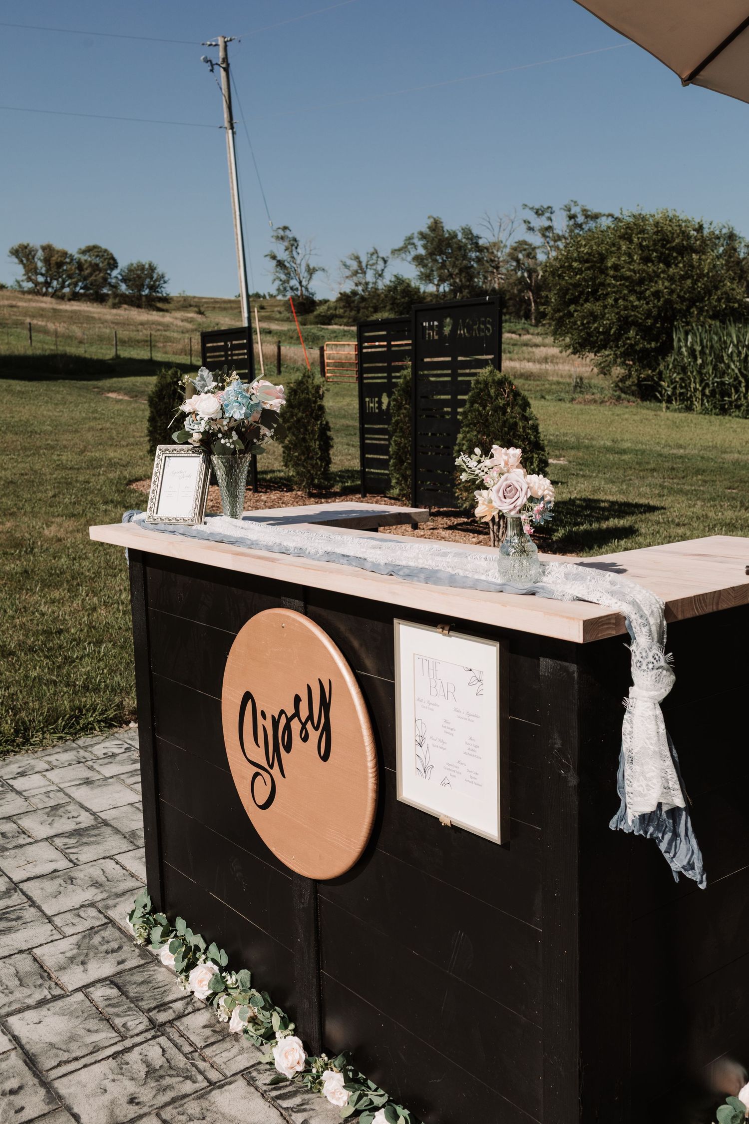 Outdoor wedding bar setup with rustic black Sipsy bar cart decorated with pink roses and blue ribbon.