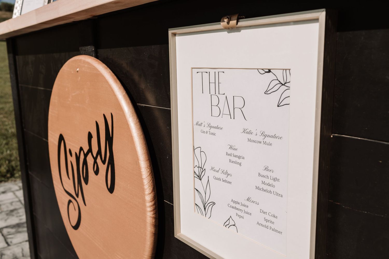 Close-up of elegant wedding bar menu sign with floral details displayed on a rustic black bar cart.