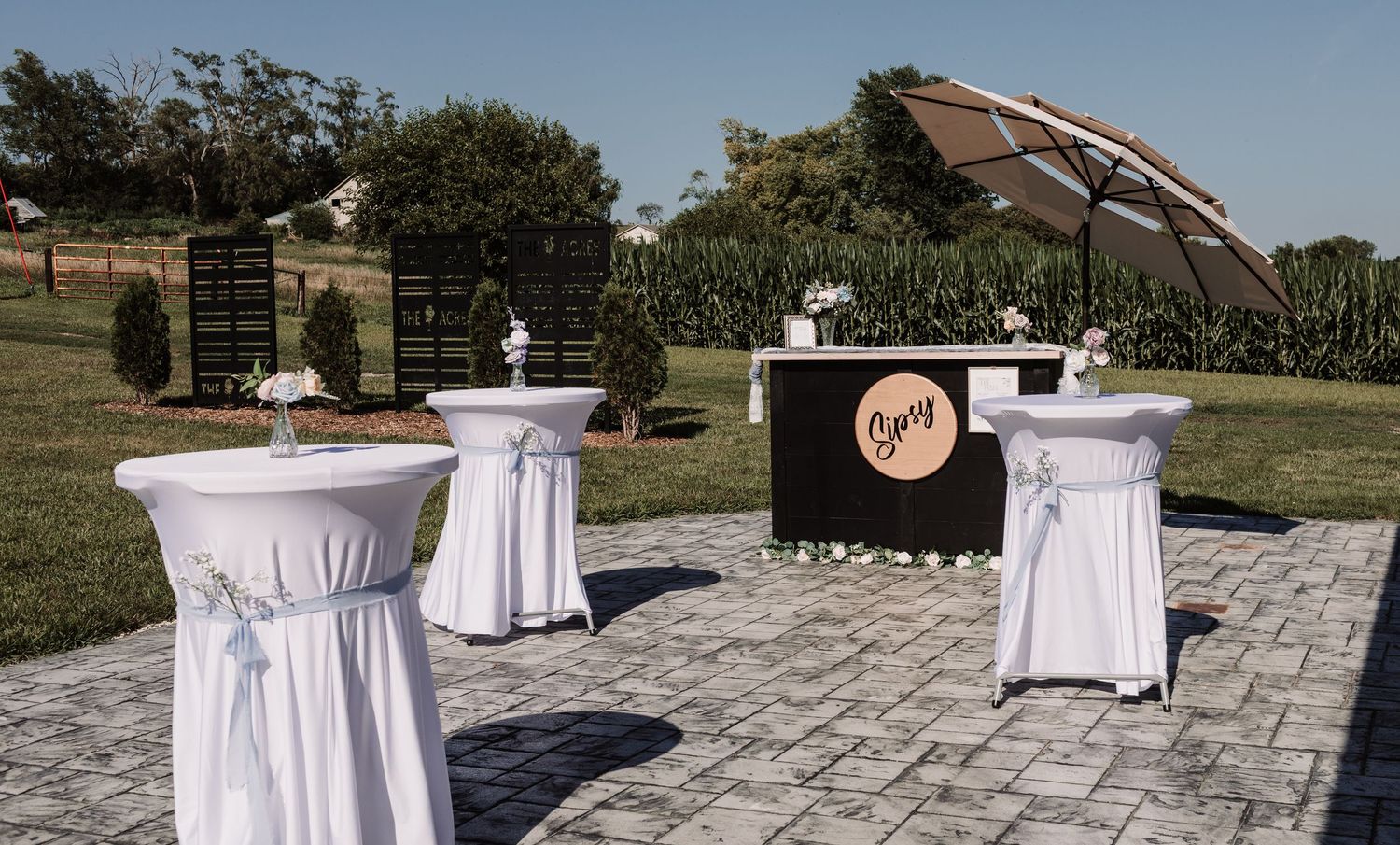 Outdoor wedding cocktail hour setup with white draped high-top tables and Sipsy bar under umbrella.