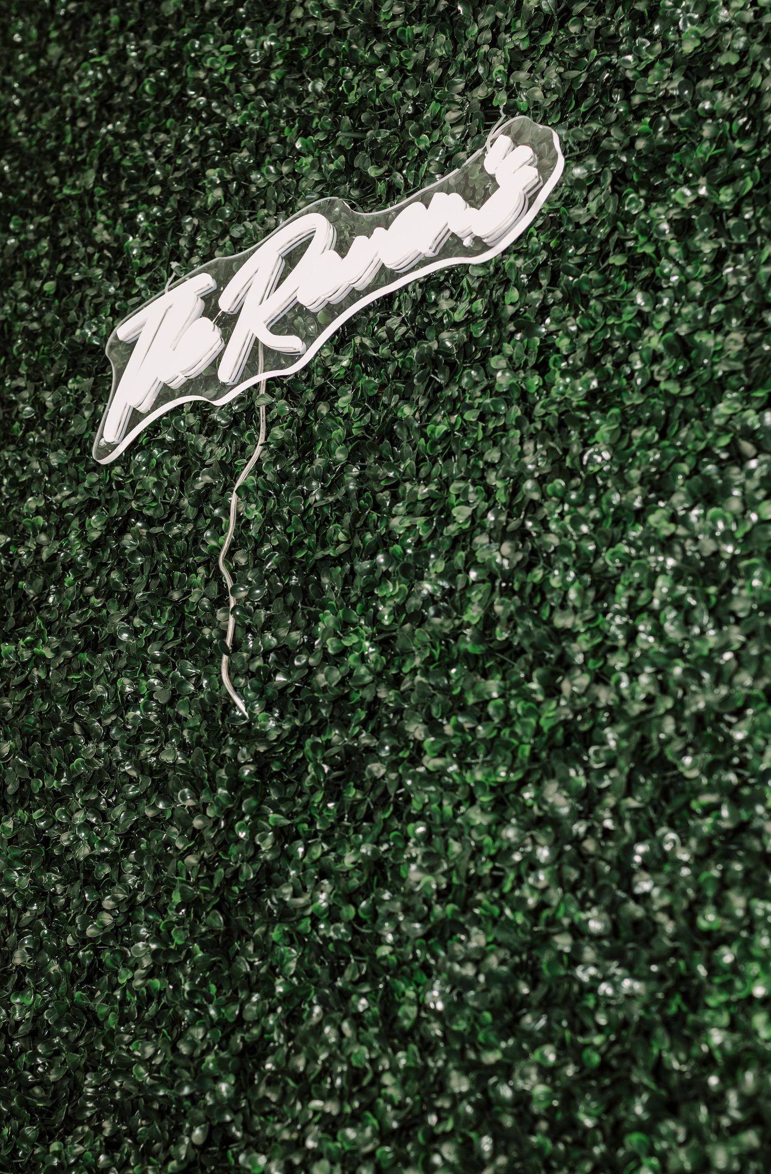 White neon cursive sign mounted on a lush green artificial hedge wall backdrop.