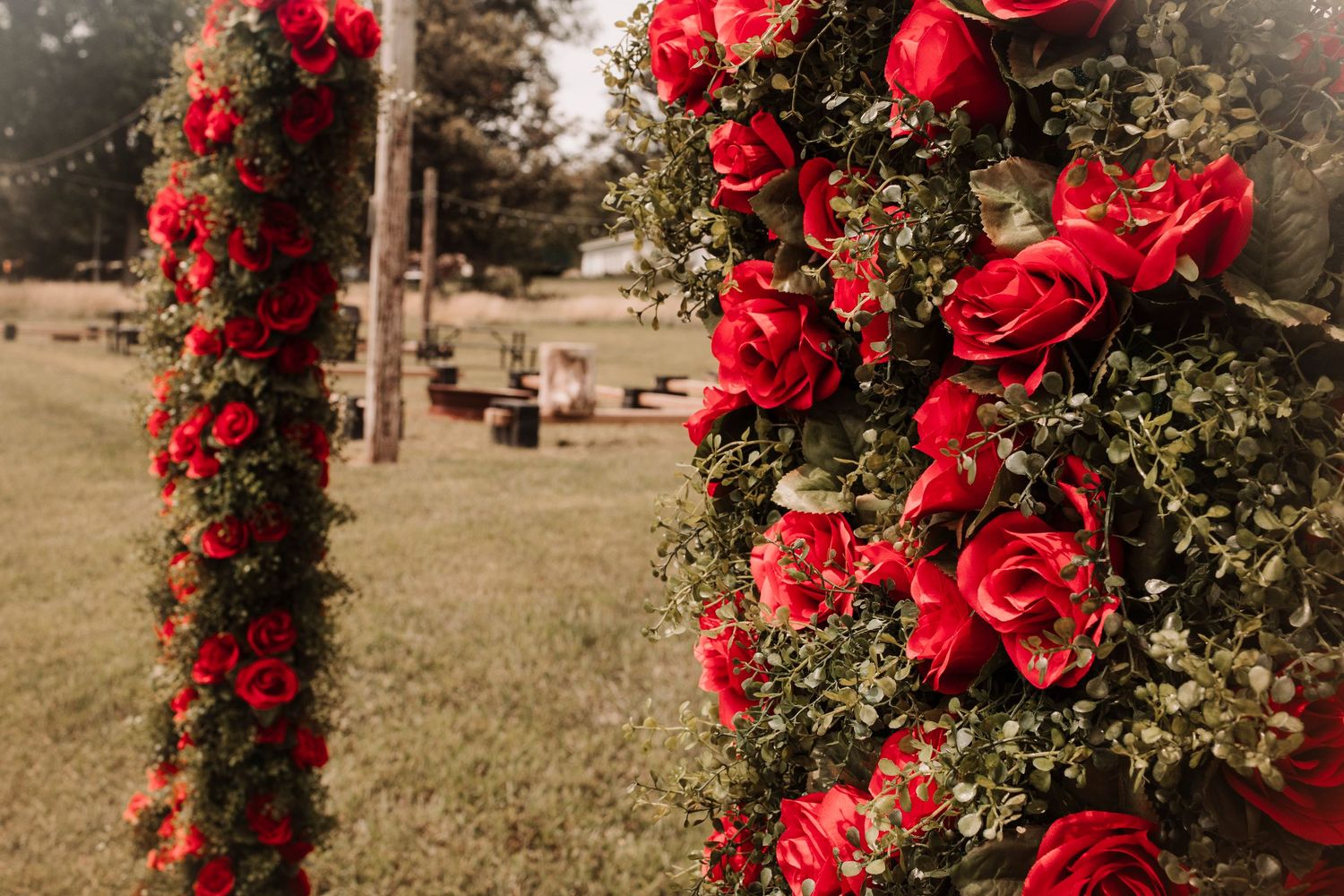 Tall floral columns covered in red roses and greenery at an outdoor wedding ceremony.
