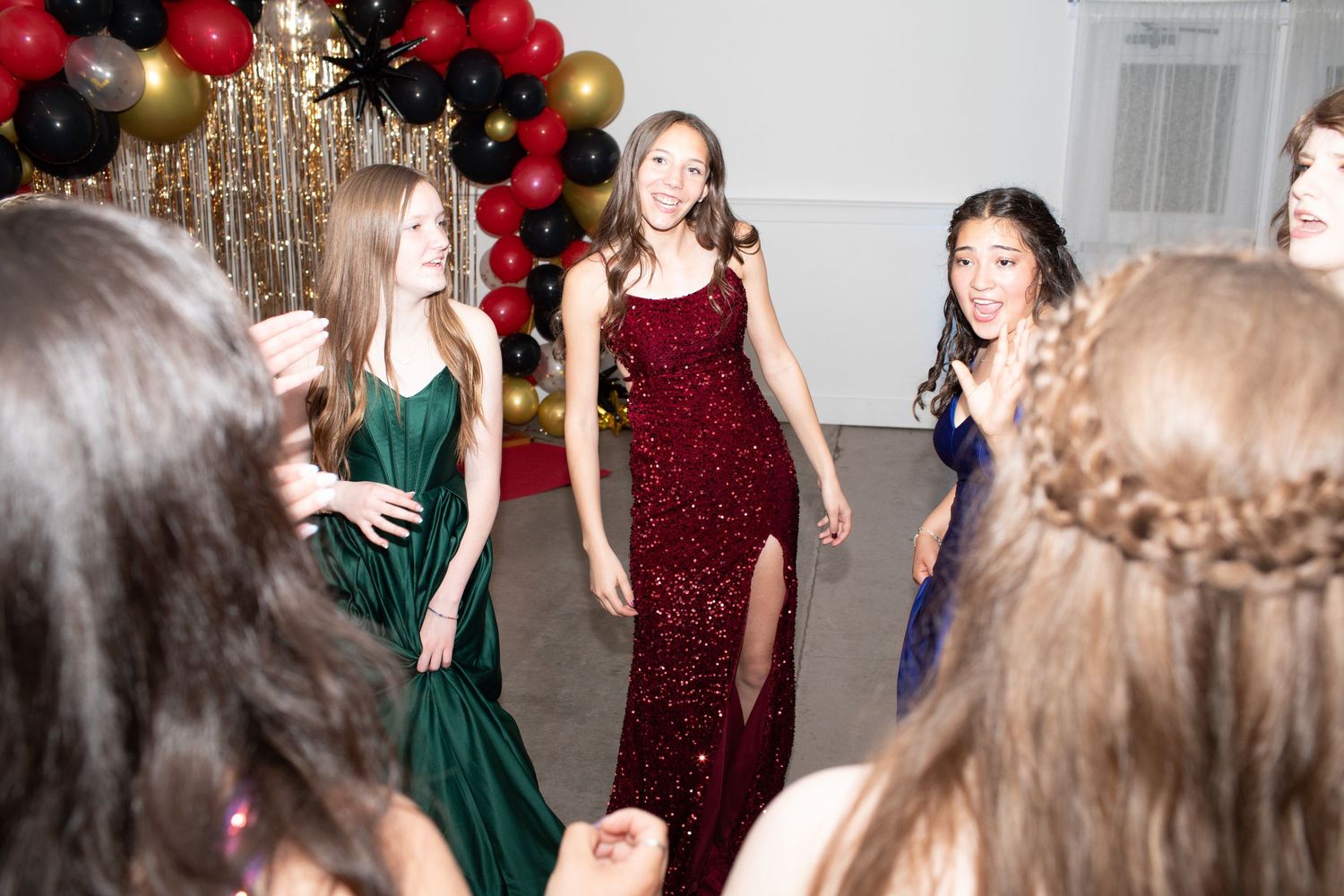Teenage girls in colorful formal gowns dancing joyfully at a prom with balloon decorations.