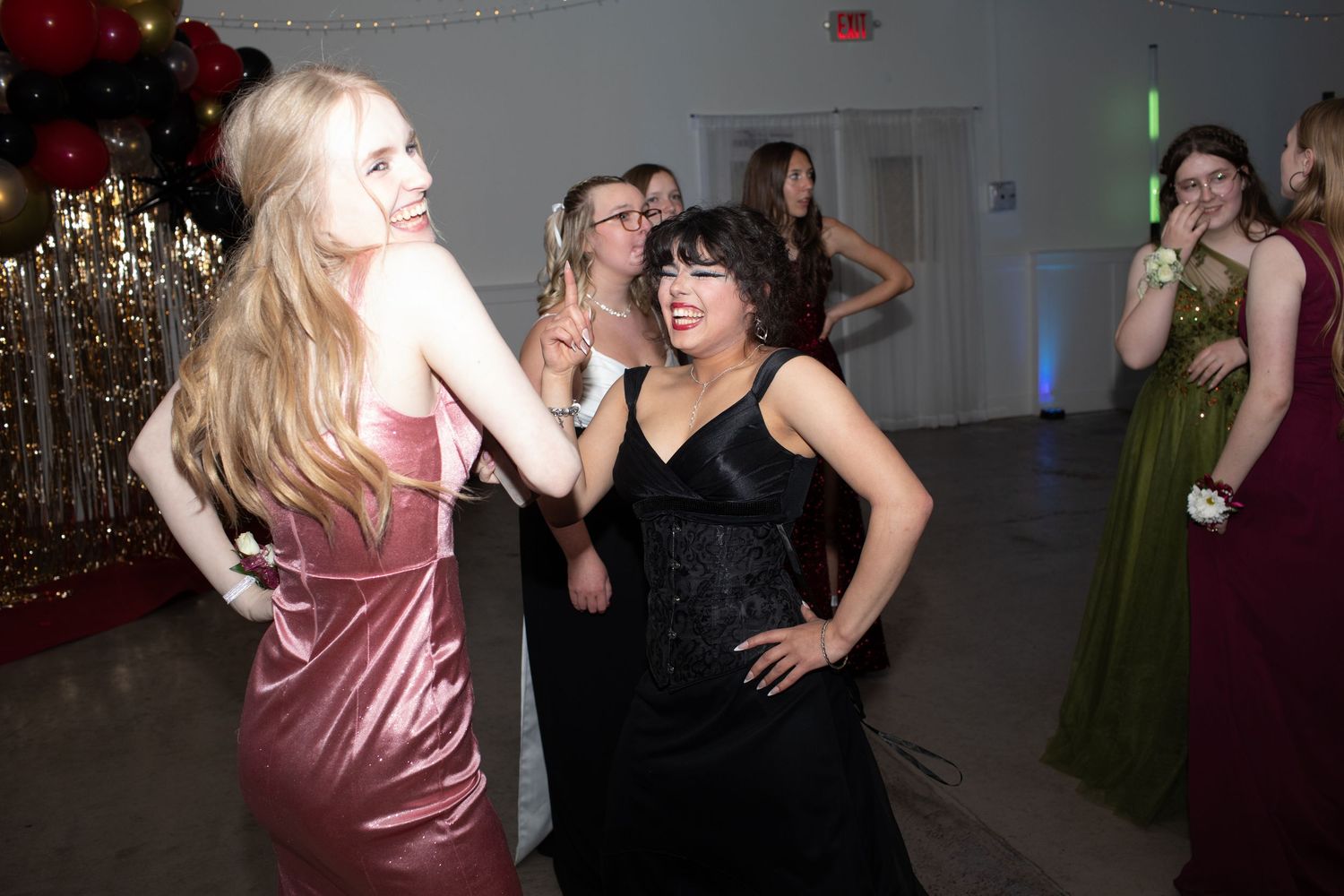 Two girls in formal dresses dancing together at a prom with gold and red balloon decor.