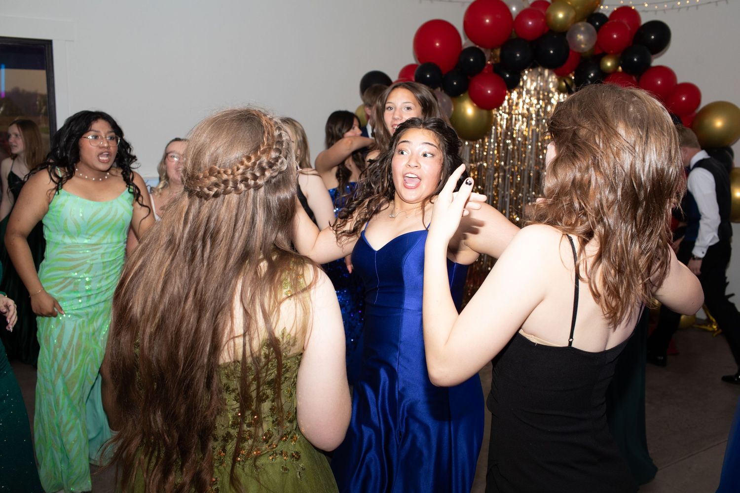 Group of teenage girls in formal gowns socializing and dancing at a decorated prom event.