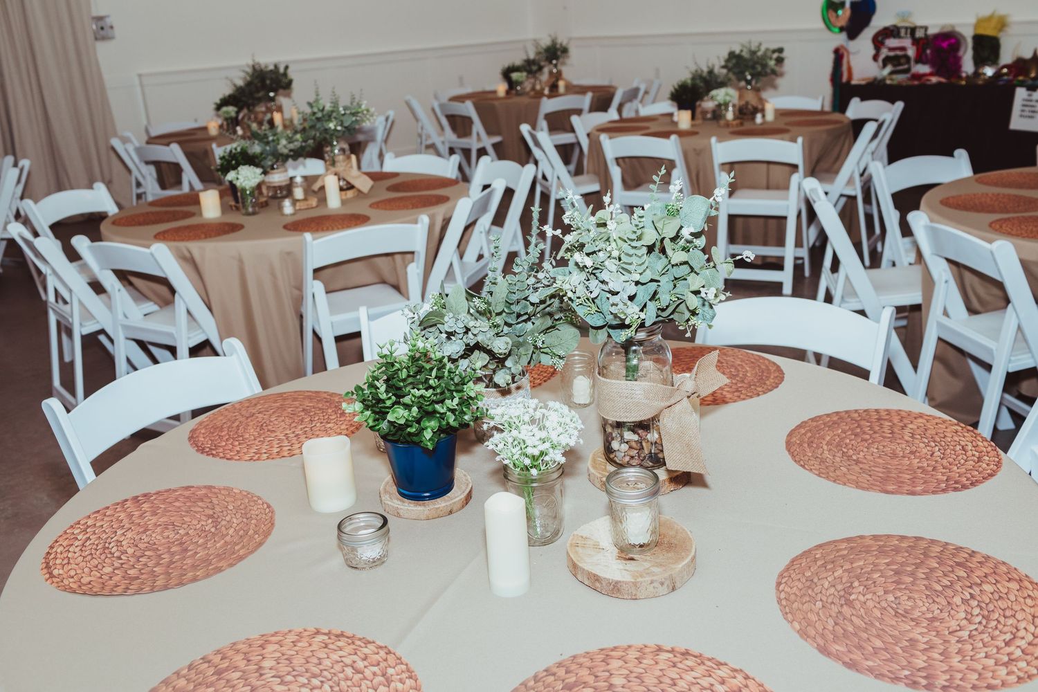 Elegant wedding reception tables with white chairs, woven placemats, and greenery centerpieces.
