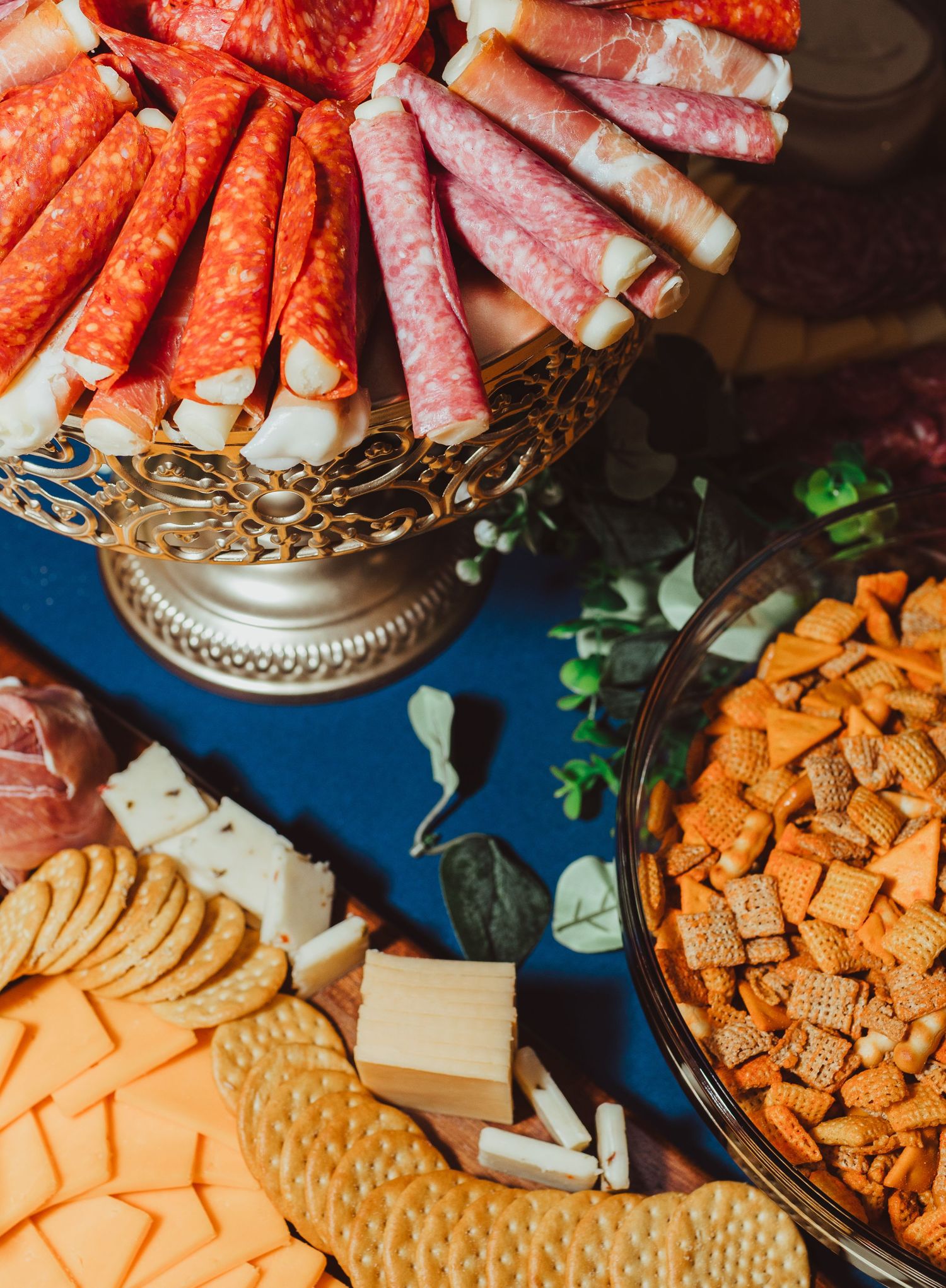 Elegant charcuterie board with rolled salami, cheese, crackers, and Chex mix on a blue tablecloth.