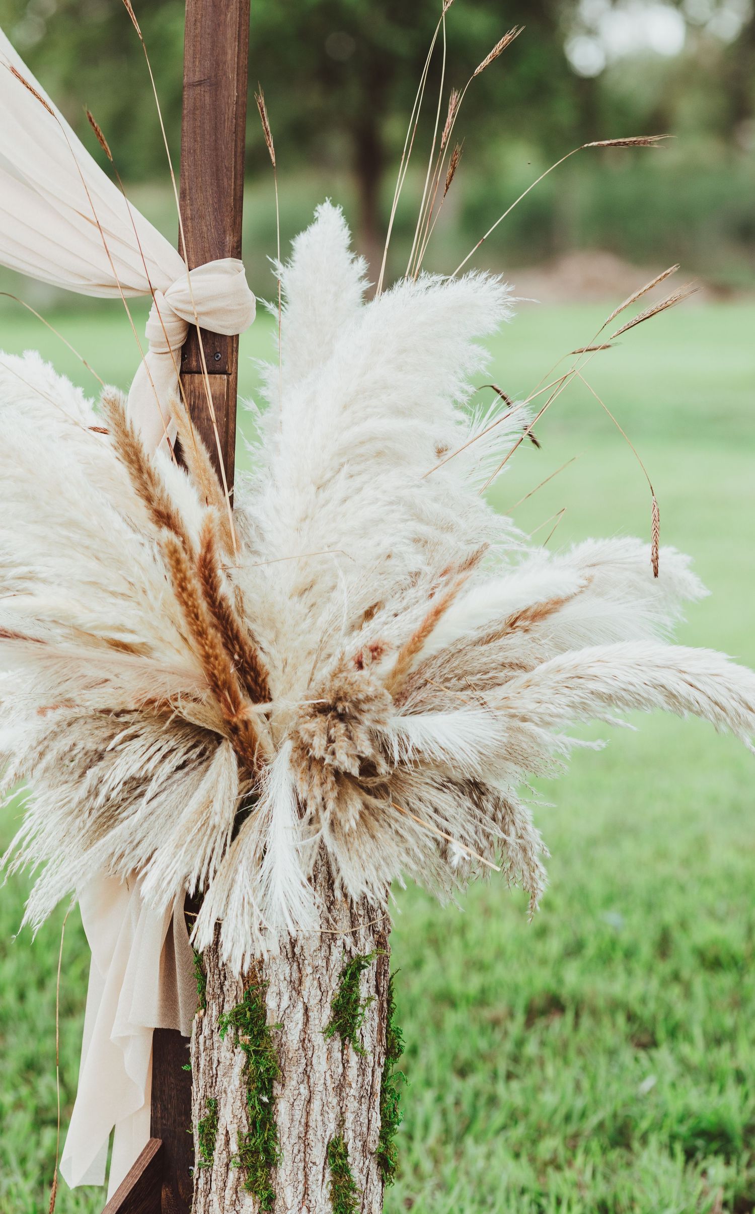 Fluffy white pampas grass bouquet tied to rustic wooden post with moss at outdoor wedding.