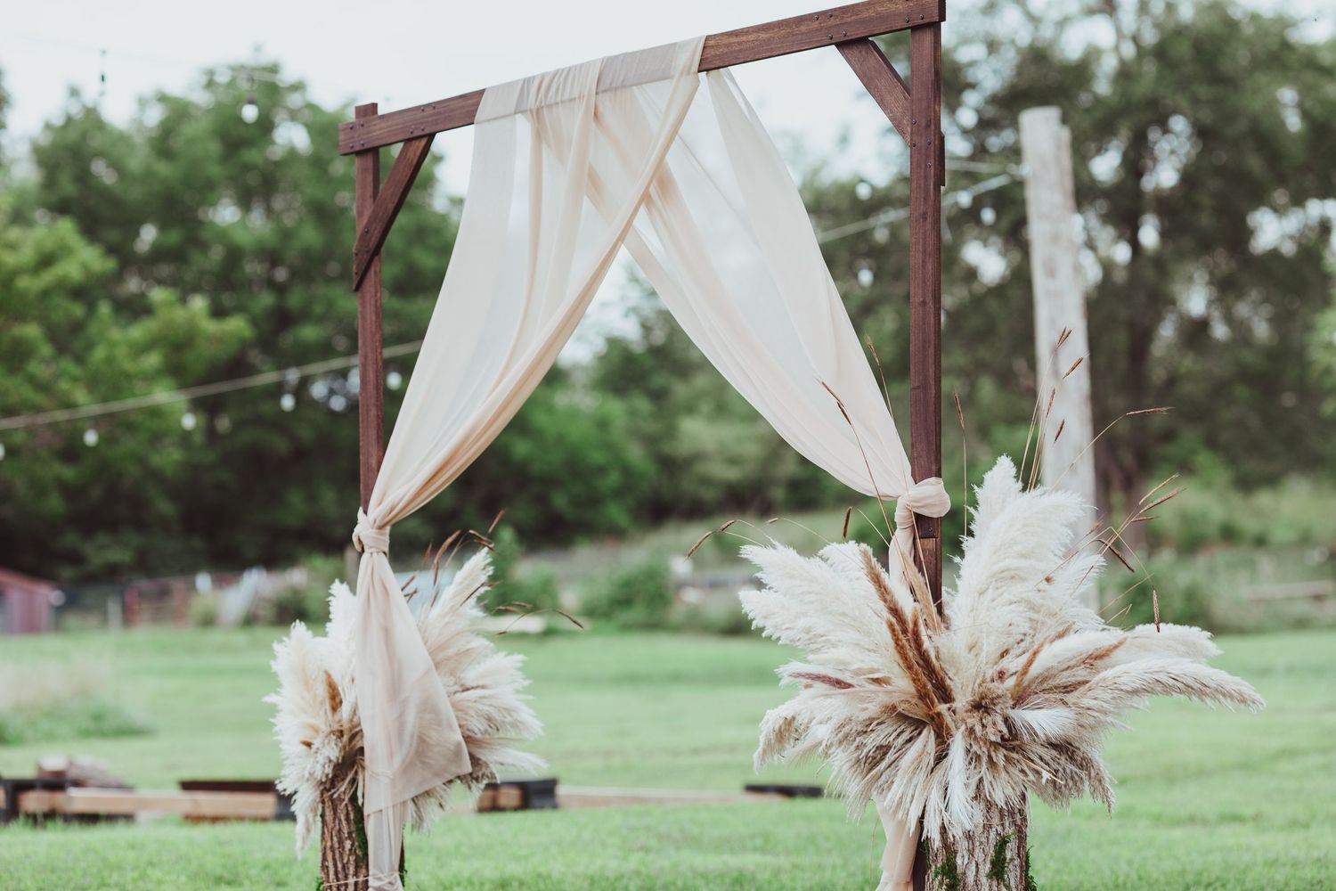 Rustic wooden wedding arch draped with white fabric and pampas grass in outdoor green field.