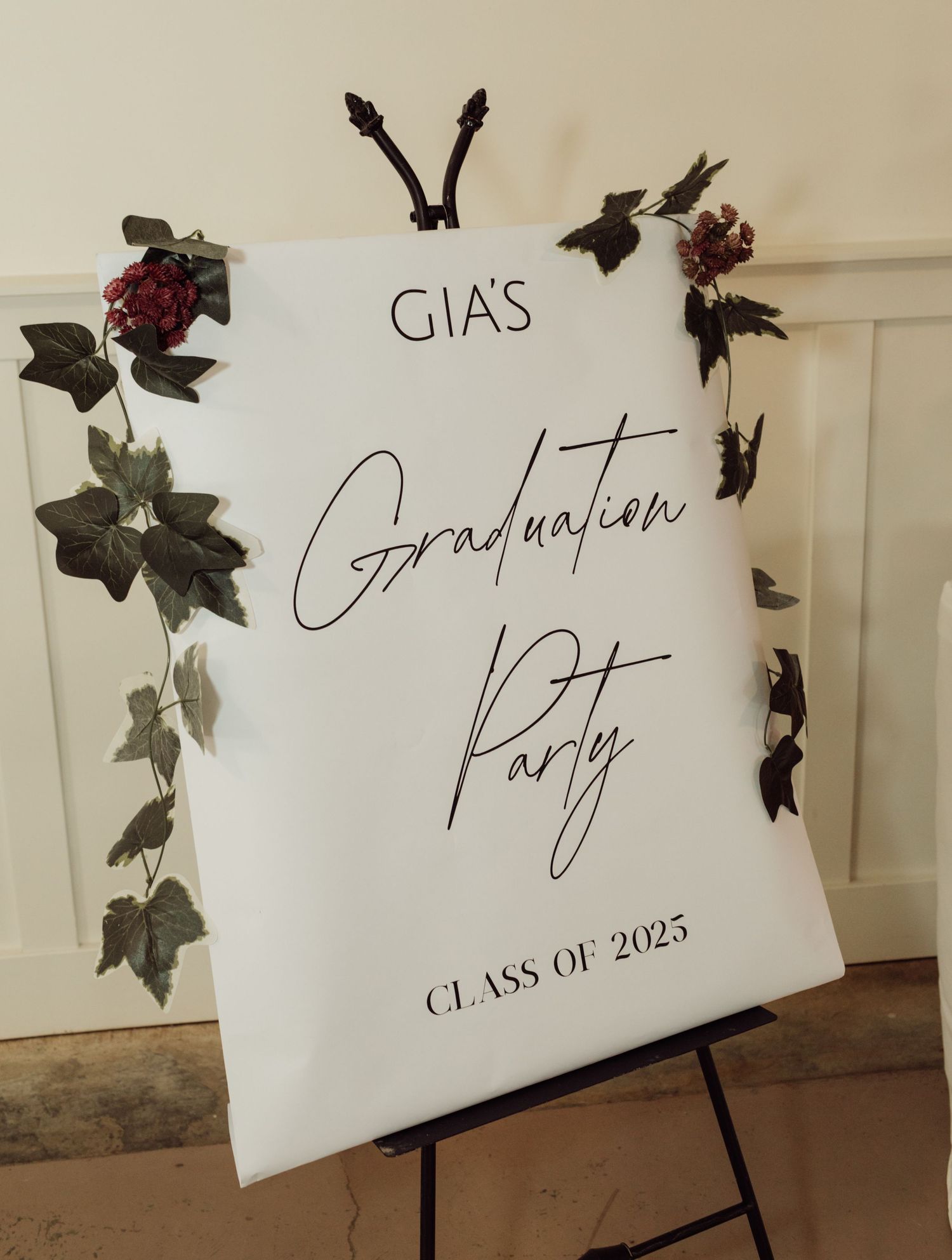 White welcome sign on easel reading Gia's Graduation Party Class of 2025 decorated with ivy and berries.