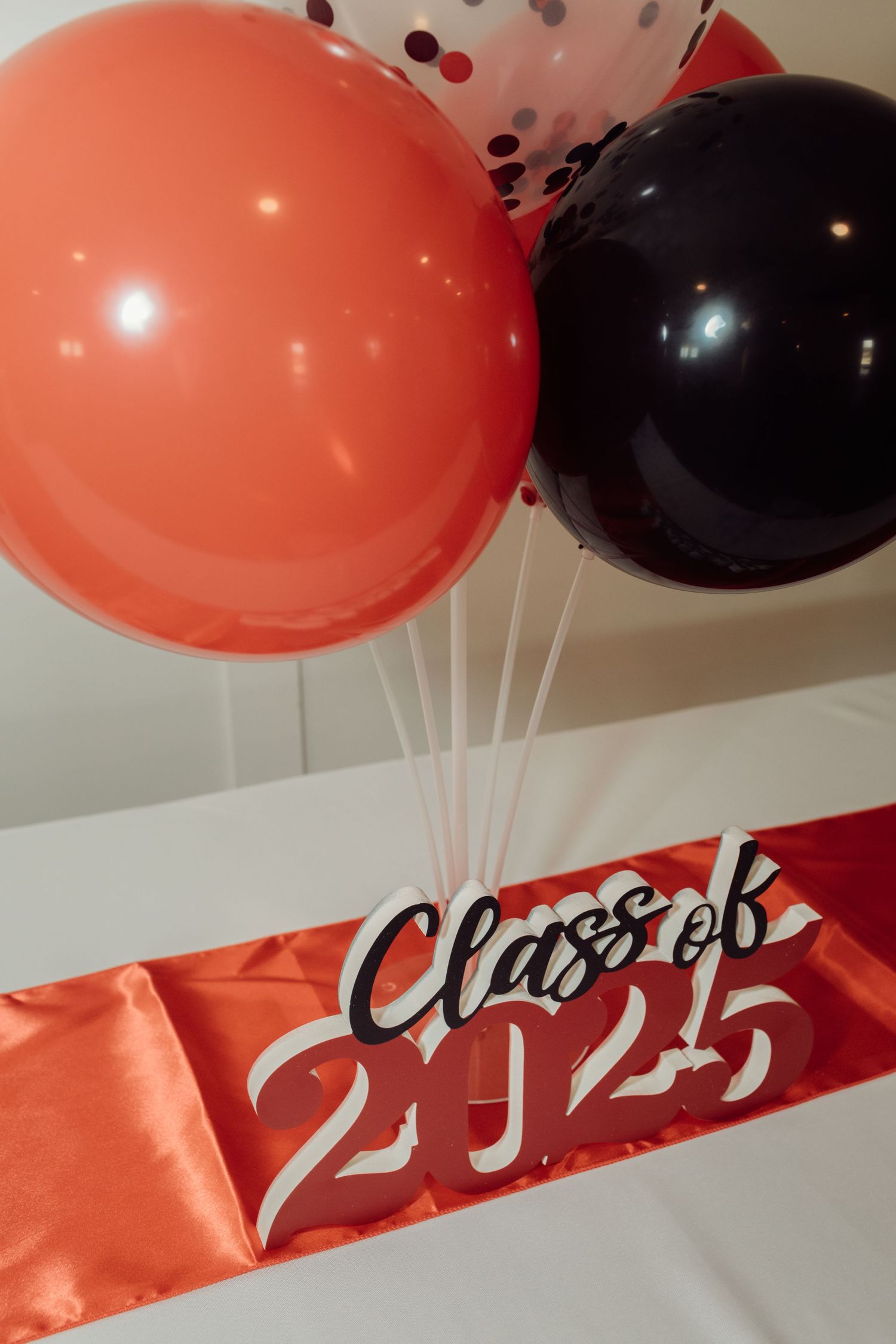 Class of 2025 graduation sash displayed with red, black, and confetti balloons on a white surface.