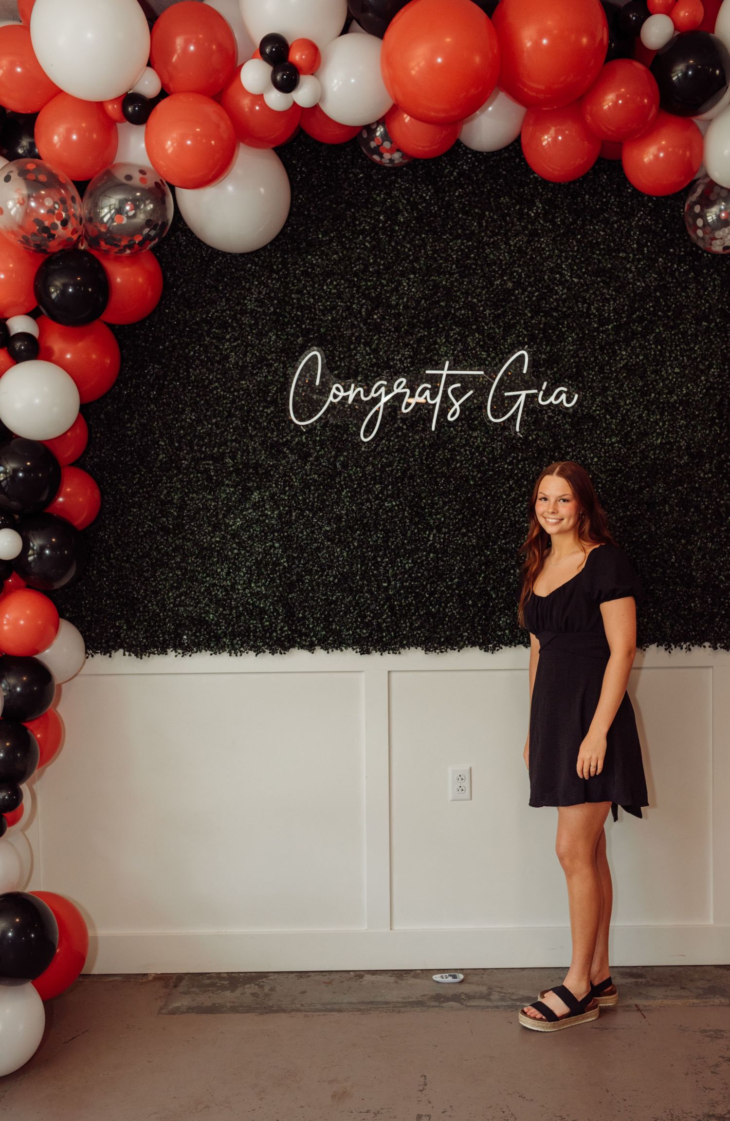 Young woman in black dress smiling in front of hedge wall with red, black, and white balloon arch and Congrats Gia neon sign.