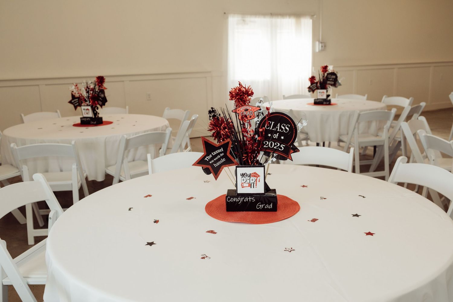 Round banquet tables with white linens and red and black Class of 2025 graduation centerpieces in a hall.