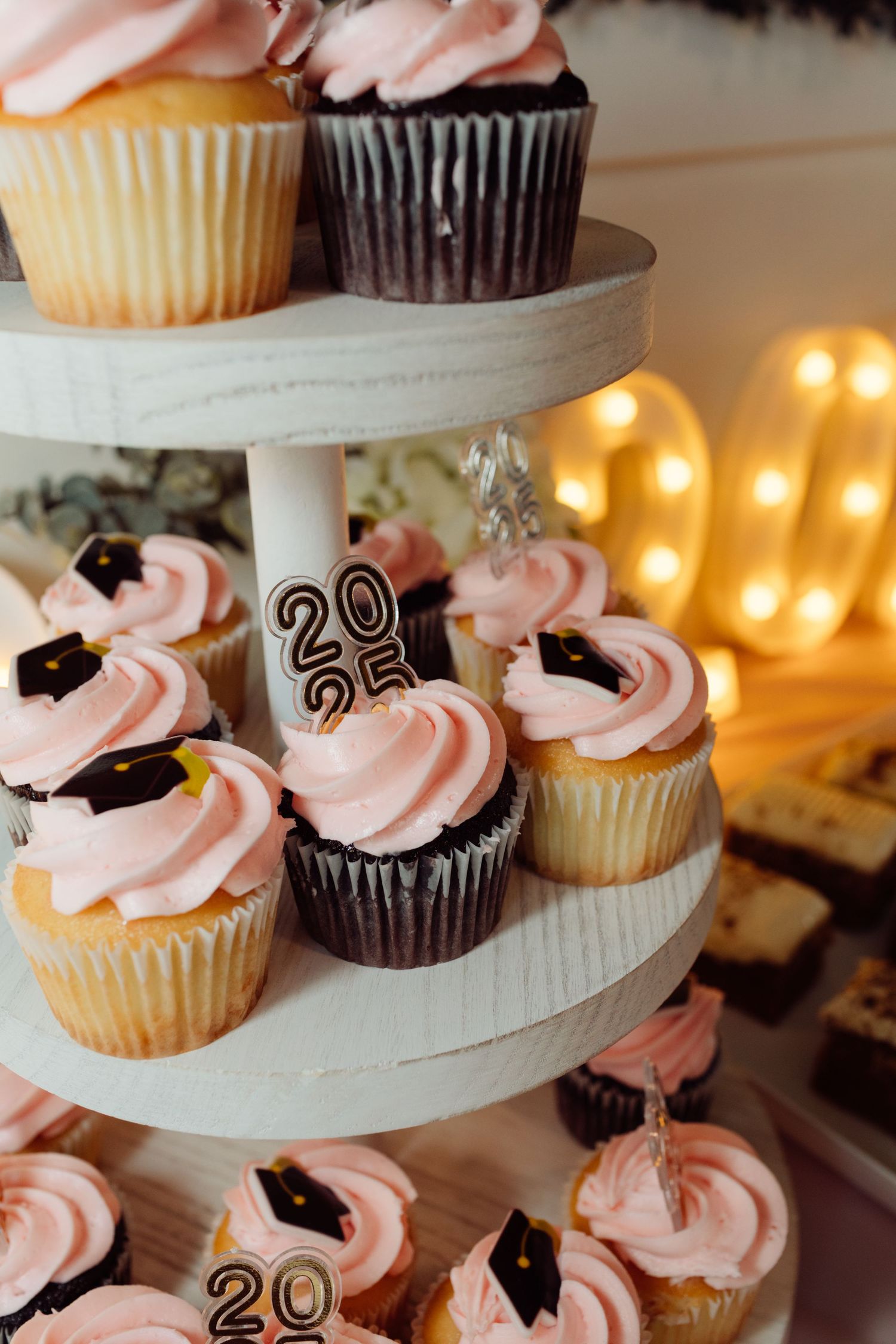 Close-up of pink frosted graduation cupcakes with 2025 and cap toppers on a white tiered stand.