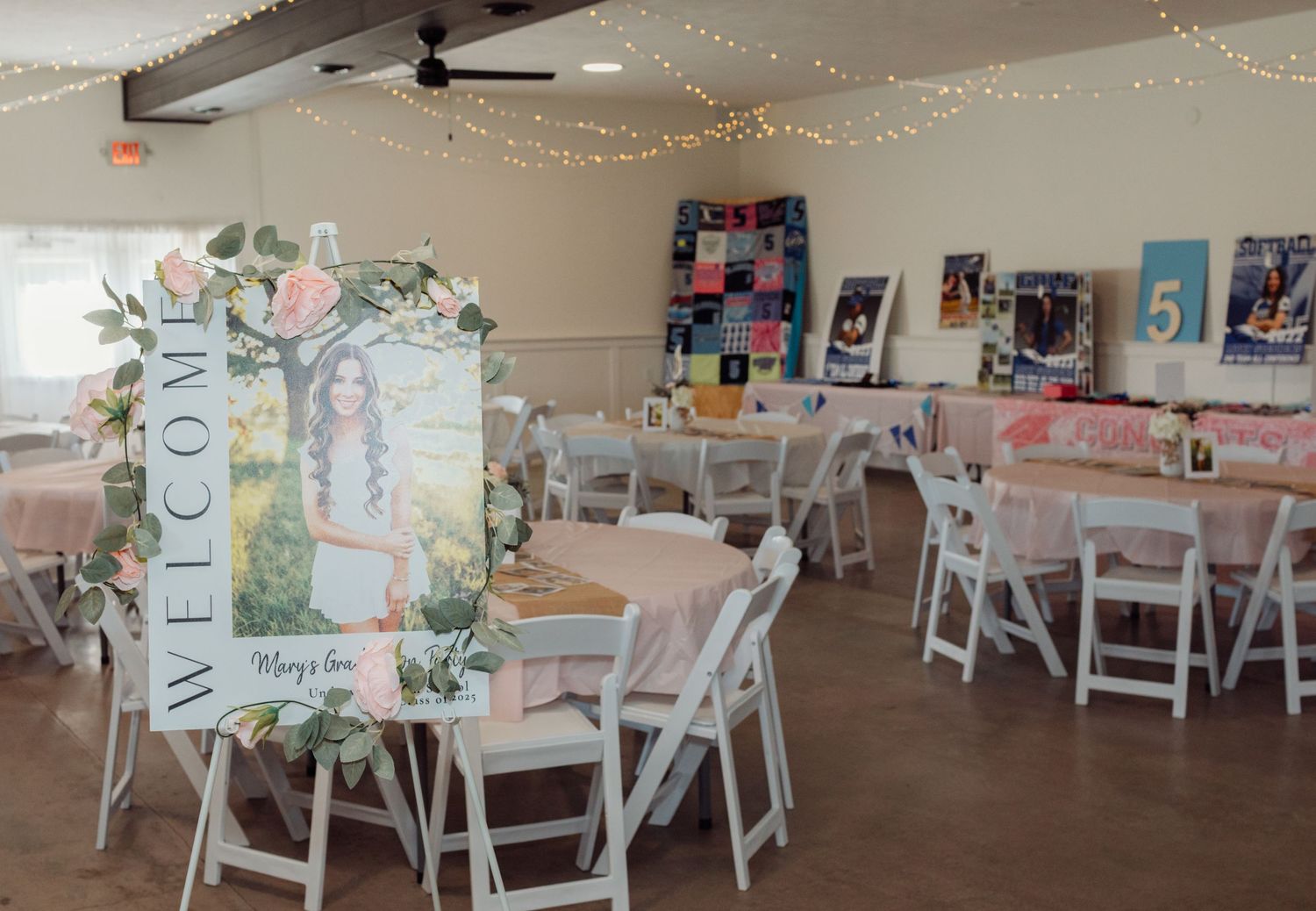 Decorated graduation party hall with welcome sign, fairy lights, white chairs, and round draped tables.