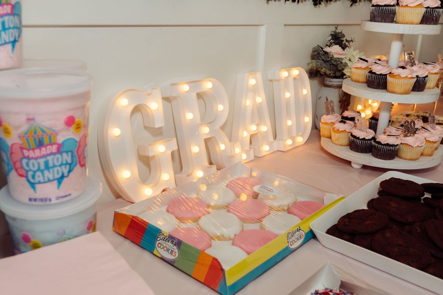 Illuminated GRAD marquee letters beside cotton candy and colorful cupcakes on a graduation dessert table.