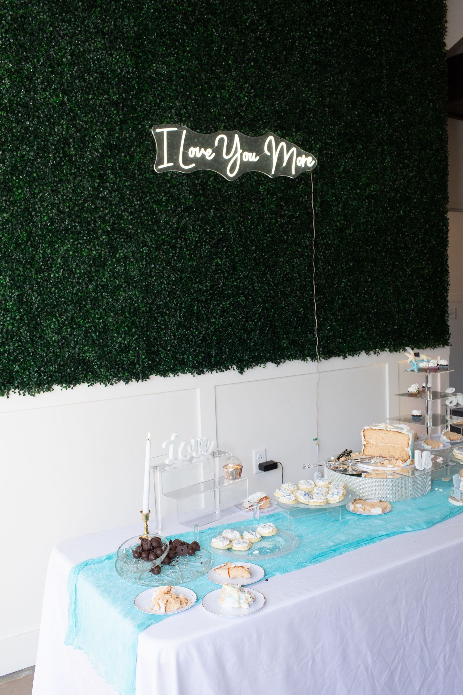 Wedding dessert table with neon I Love You More sign on green hedge wall backdrop.