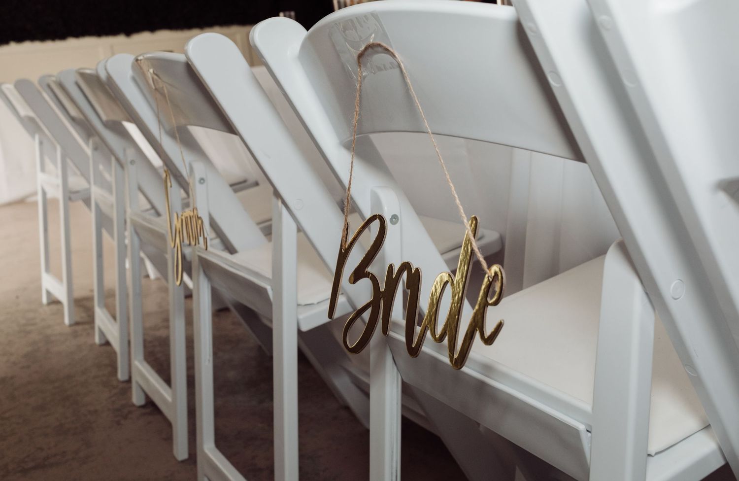 White folding chairs with gold Bride wooden sign hanging at wedding ceremony setup.