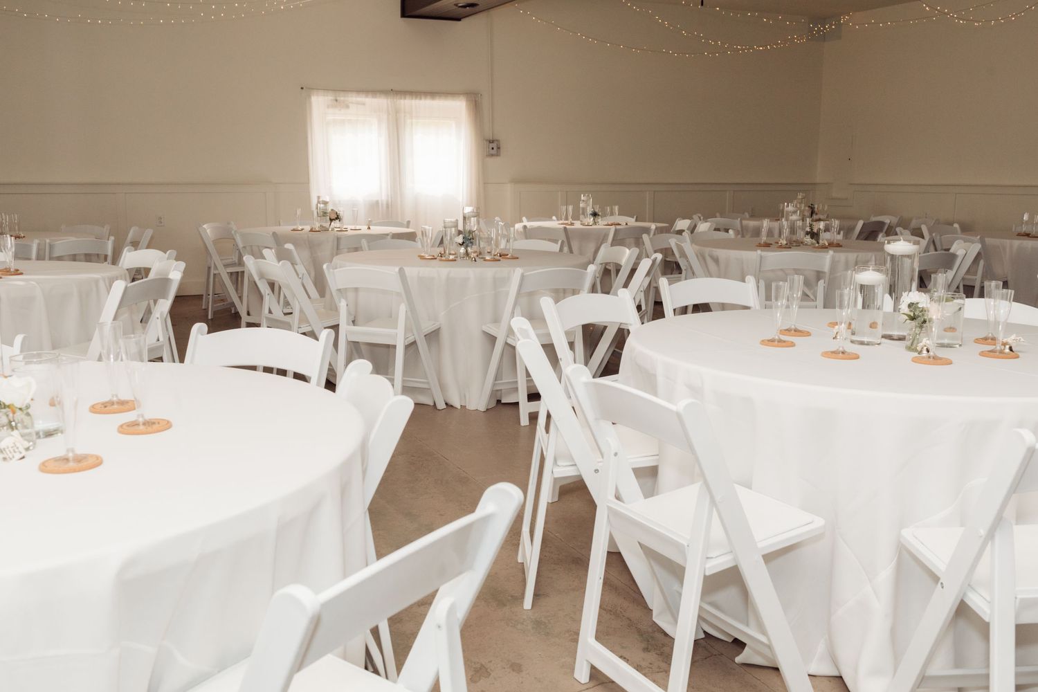Elegant wedding reception hall with round tables draped in white linens, white folding chairs, and soft natural lighting.