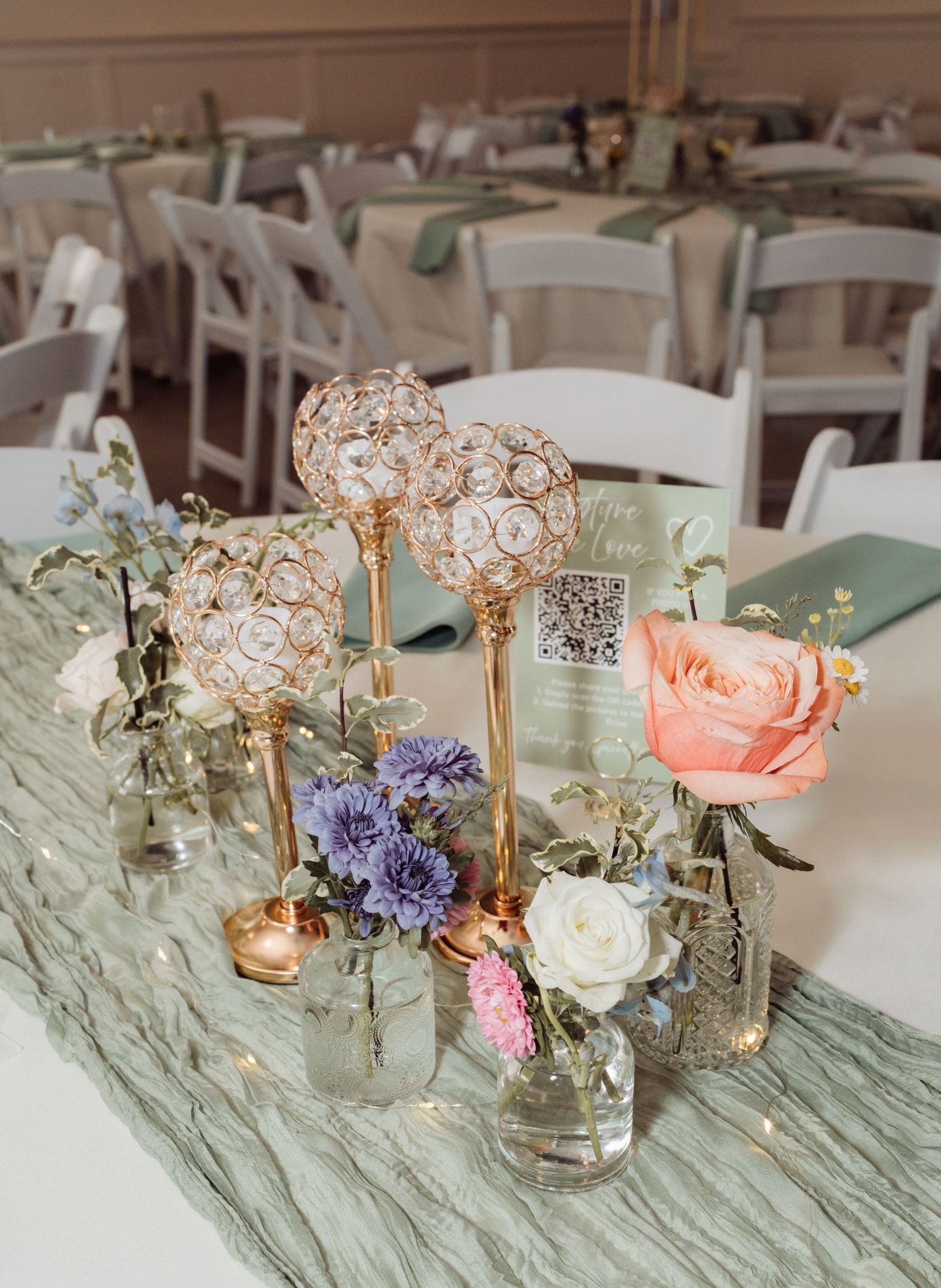Wedding table centerpiece with gold candelabras, crystal globes, and colorful floral arrangements on sage green runner.