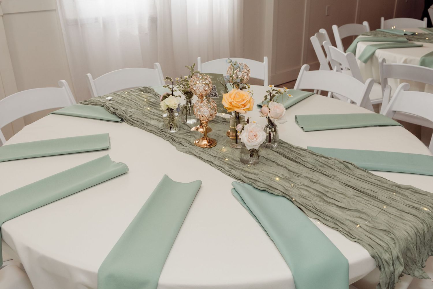 Round wedding reception table with white linen, sage green napkins, textured runner, and floral centerpiece.