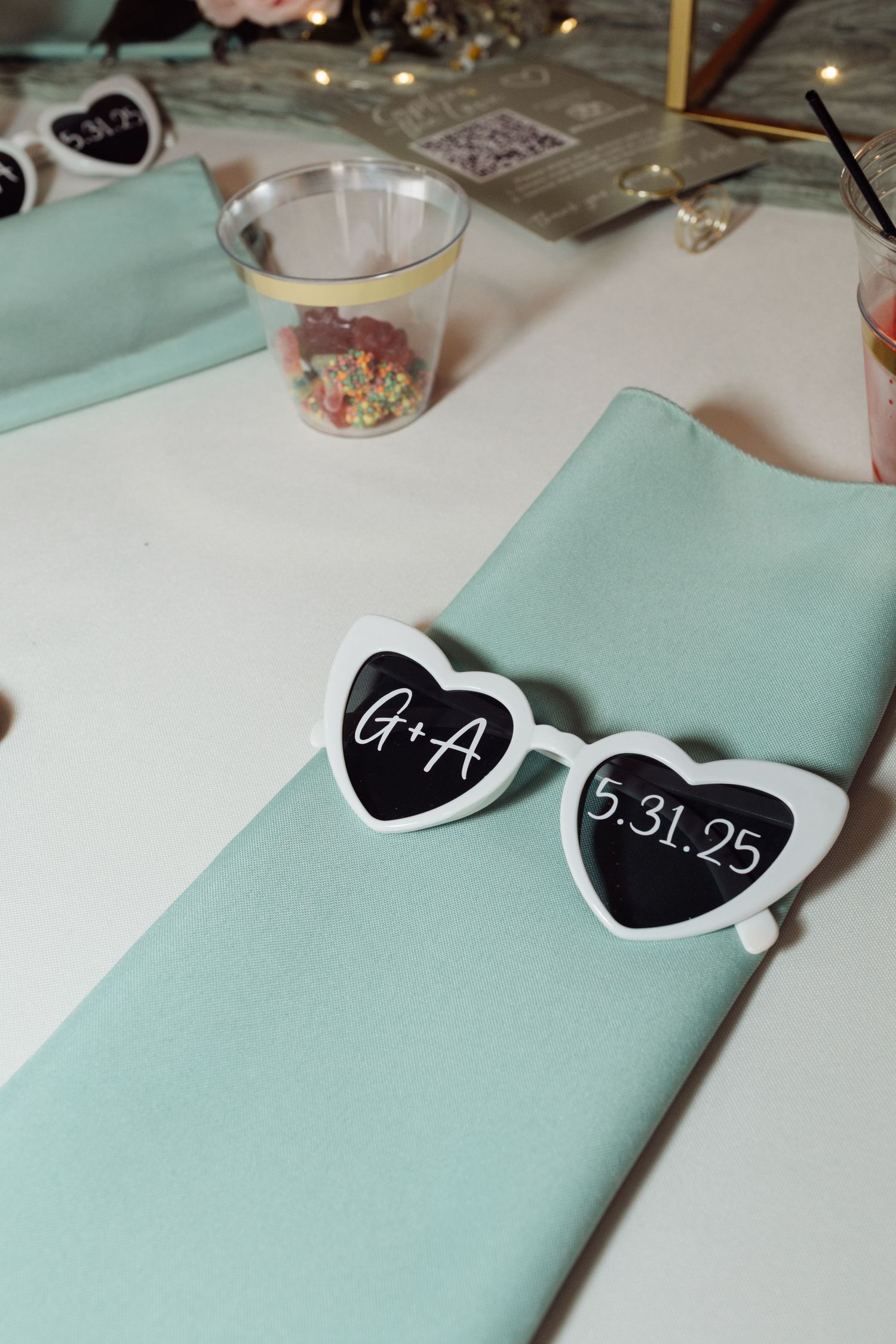 Heart-shaped sunglasses with G+A and 5.31.25 wedding date displayed on a mint green napkin at reception table.